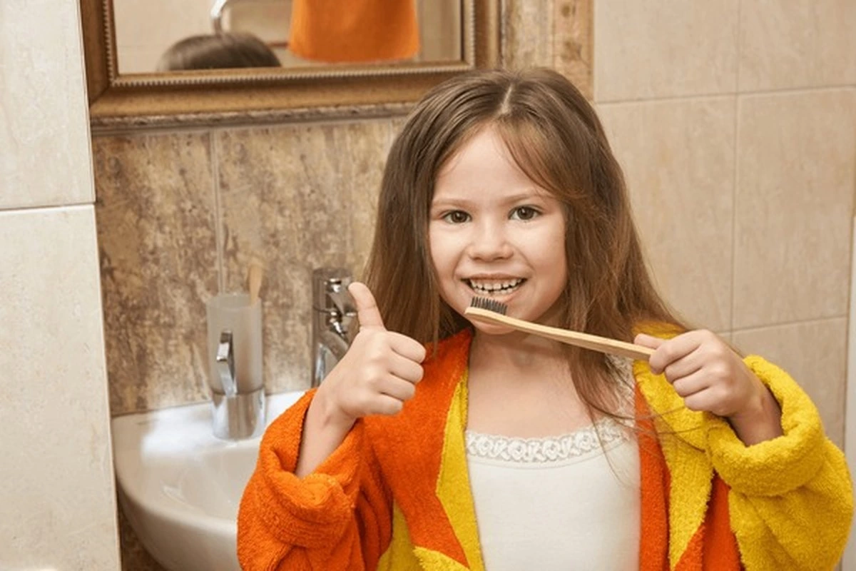 Pediatric pulpotomies Frisco TX: girl thumbs-up brushing teeth with wooden brush for healthy smile at Dental Smart Kids
